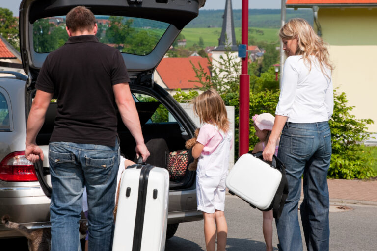 Family loading a car trunk on a sunny day: man and woman with two young girls and luggage by the silver hatchback.