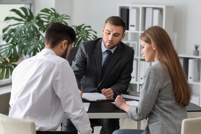 Three coworkers gather around a desk, examining documents; a man in a dark suit leads the discussion while a woman in a checkered blazer and another man listen.