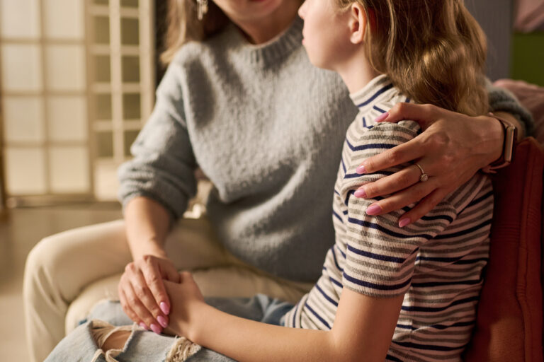Caucasian mother comforting teenage daughter while holding hands and embracing shoulder during serious home conversation, representing parental support. Useful for family counseling campaigns